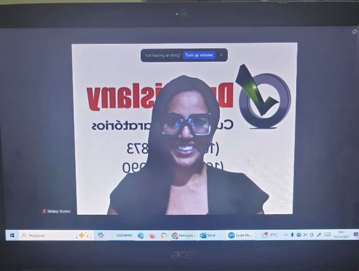 Dra Gislany sorrindo em aula on-line ao vivo, com o fundo virtual do Dra Gislany Cursos Preparatórios na tela do notebook