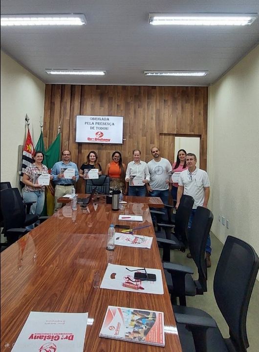 Sala de reunião com mesa comprida e participantes ao fundo, diante do slide de encerramento