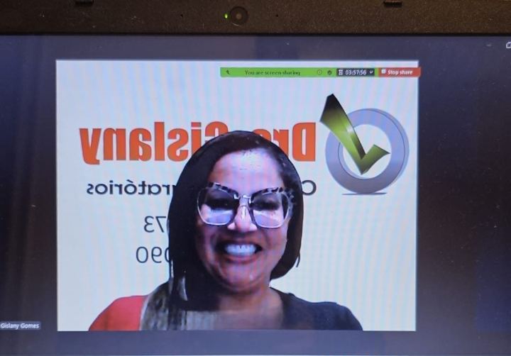 Dra Gislany de óculos sorrindo durante aula on-line, com a logomarca do Dra Gislany Cursos Preparatórios ao fundo