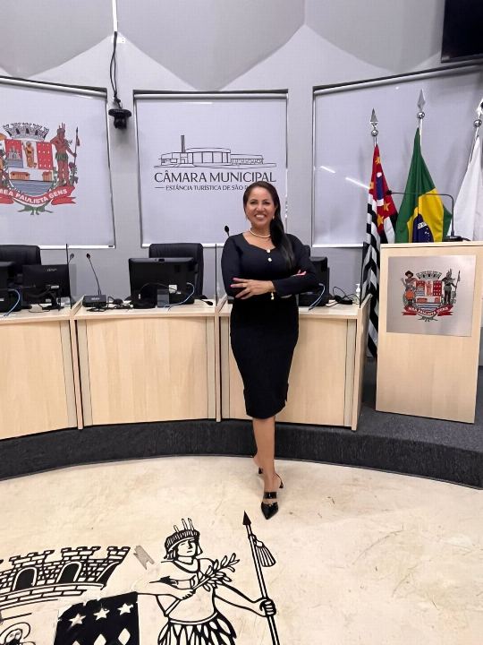 Dra Gislany em pé no plenário da câmara municipal, diante da mesa diretora e das bandeiras oficiais