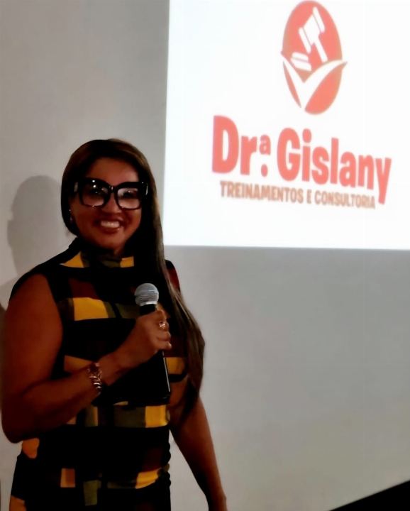 Dra Gislany ao microfone diante do painel com a marca Dra Gislany Treinamentos e Consultoria